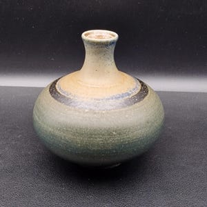 Könnte beinhalten: Eine handgefertigte Keramikvase mit rundem Körper und schmalem Hals. Die Vase hat eine Farbpalette aus gedämpftem Grün, Beige und Blau, mit einem dunklen Band, das die Mitte umgibt. Die Vase eignet sich zum Präsentieren von Blumen oder als Dekorationsartikel.