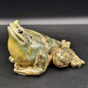 Op de afbeelding: Een keramieken kikkerbeeldje in groen, geel en bruin. De kikker zit met open mond. Het beeldje heeft een getextureerd oppervlak en gedetailleerde kenmerken.