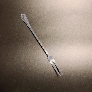 May include: A silver fork with a rounded handle and two prongs. The fork is lying on a gray surface.