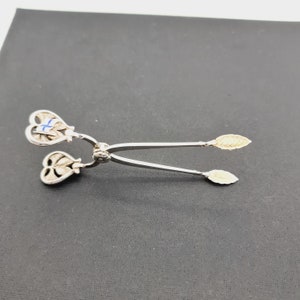 Op de afbeelding: Zilveren suikertang met hartvormige handvatten. Een handvat heeft een blauw en wit emaille ontwerp, terwijl het andere handvat een bladontwerp heeft. De tang heeft goudkleurige uiteinden.