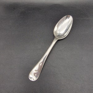 May include: A silver spoon with a rounded bowl and a long, slender handle. The spoon is highly polished, reflecting the light. The handle has a decorative element near the bowl. The spoon is set against a dark background.