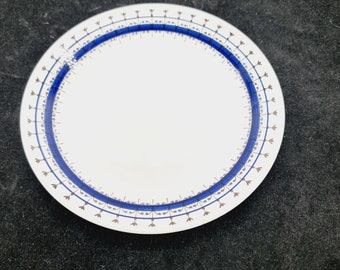 May include: A white ceramic plate with a blue and brown decorative border. The border features a repeating pattern of small blue flowers and brown lines.