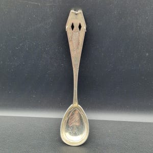May include: A silver spoon with an ornate design. The handle features a fish and two diamond-shaped cutouts. The spoon's bowl has an etched design. The spoon is set against a dark background.