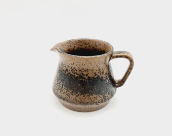 May include: A small brown ceramic creamer with black speckles and a wide, rounded base. The creamer has a handle on the right side.