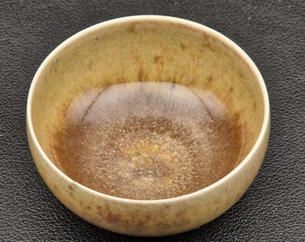May include: A small, round ceramic bowl with a speckled, glazed interior. The bowl's interior transitions from a dark brown at the center to a lighter, golden-brown hue towards the rim. The exterior is a muted, yellowish-brown.