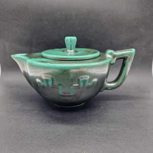 Könnte beinhalten: Eine türkisfarbene Keramik-Teekanne mit passendem Deckel und Griff. Die Teekanne hat einen abgerundeten Körper mit Ausguss und Griff. Der Deckel hat einen kleinen, vertikalen Griff. Die Teekanne hat eine dunkle, strukturierte Oberfläche mit einem geometrischen Design.