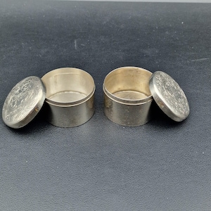 Può includere: Due piccoli contenitori rotondi color argento con coperchi. I coperchi sono leggermente socchiusi, rivelando l'interno dei contenitori. Ogni coperchio presenta un motivo floreale in rilievo. I contenitori sono su uno sfondo scuro.