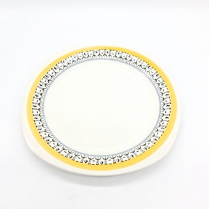 May include: A white ceramic dinner plate with a yellow and black patterned border. The pattern features a repeating design of stylised trees.