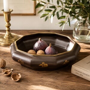 May include: A dark brown octagonal ceramic bowl with a gold-toned rim and gold leaf-shaped accents on the exterior. The bowl contains two fresh purple figs and two whole walnuts, with additional walnut shells scattered nearby on the surface.