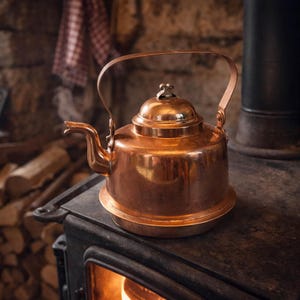 Könnte beinhalten: Ein polierter Kupferkessel mit gebogenem Griff und Ausguss steht auf einem schwarzen Ofen. Der Kessel hat einen runden Boden und einen Deckel mit einem kleinen dekorativen Knopf. Die warmen Kupfertöne kontrastieren mit dem dunklen Ofen und erzeugen eine gemütliche, rustikale Atmosphäre.