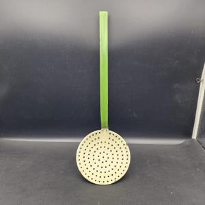 Könnte beinhalten: Ein Vintage-Küchenschöpfer mit rundem, perforiertem Kopf und langem, grünem Griff. Der Kopf des Schöpfers ist cremefarben mit zahlreichen kleinen Löchern. Der Griff ist einfarbig hellgrün. Dieses Utensil dient zum Abseihen von Flüssigkeiten oder zum Entfernen von Feststoffen.