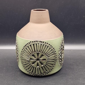 Może przedstawiać: Ceramiczny wazon z jasnobrązową górą i szałwiowym zielonym korpusem. Korpus ma powtarzający się okrągły wzór w ciemniejszym, metalicznym kolorze. Wazon ma zaokrąglony kształt i wąski otwór.