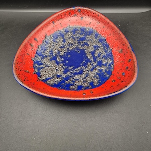 May include: A triangular ceramic dish with a vibrant color scheme. The outer rim is a bold red, transitioning to a deep blue center. The blue area has a textured, speckled appearance. The dish is likely for decorative or serving purposes.