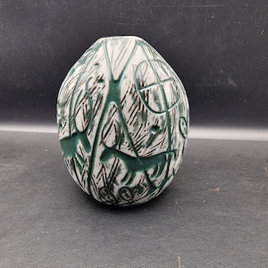 May include: An oval-shaped ceramic vase with a white base, adorned with green and black abstract patterns. The vase features carved designs and animal-like figures. A decorative item for the home.