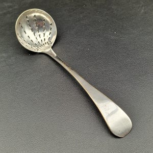May include: A silver-coloured, slotted spoon with a long handle. The spoon's bowl is round with numerous small holes. The handle has a smooth, curved design. The spoon is likely used for straining or serving.