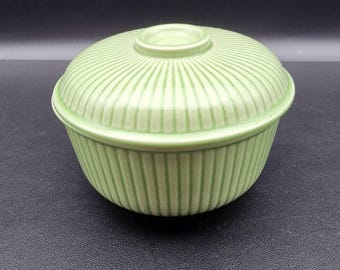 May include: A light green ceramic bowl with a matching lid. The bowl and lid have vertical ribbed detailing. The lid has a small, circular handle. The bowl is round and sits on a dark surface.