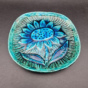 May include: A ceramic dish with a raised sunflower design. The sunflower is a vibrant blue with a textured center and petals. The dish has a turquoise rim and a textured background, creating a three-dimensional effect. The overall design is artistic and decorative.
