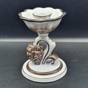 May include: A white ceramic candle holder with brown accents. The candle holder has a scalloped rim and a detailed brown design. A brown pinecone-like shape is on the stem, and the base is round.