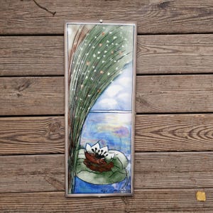 May include: Rectangular framed artwork featuring a water lily scene. The artwork depicts a water lily with a brown and white flower, a green lily pad, and a blue water background. Tall green grass and a blue sky are also visible.