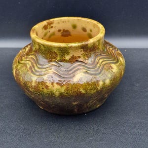 May include: A small, handcrafted ceramic vase with a rounded body and a wide opening. The vase features a textured surface with a blend of green, brown, and yellow hues. Wavy lines encircle the body, adding a decorative element.