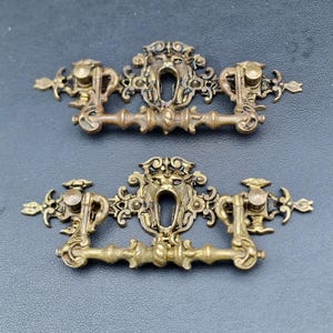 May include: Two antique brass drawer pulls with a detailed lion's head design. The pulls have ornate scrollwork and a vintage aesthetic, perfect for adding a touch of elegance to furniture. Each pull measures approximately 5 inches in length.