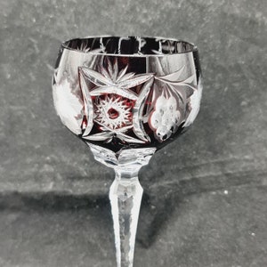 Könnte beinhalten: Ein klares Weinglas mit rotem, geschliffenem Glasdesign. Das Glas hat einen langen, schlanken Stiel und eine runde Schale.