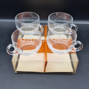 Könnte beinhalten: Set aus vier transparenten Teetassen mit Henkeln, präsentiert in einer transparenten Kunststoffbox mit einem Kartoneinsatz. Auf dem Karton steht das Wort "ORREFORS". Die Tassen sind in zwei Reihen zu je zwei angeordnet.