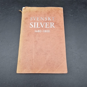 May include: A vintage book with a faded, reddish-brown cover. The title "SVENSKT SILVER" is printed in silver lettering, with the years "1680-1800" below. The book has a worn appearance, suggesting age and use.