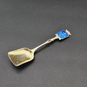 Puede incluir: Cuchara de plata con un cuenco cuadrado y un mango decorativo. El mango presenta un escudo de esmalte azul con una corona dorada. La cuchara es probablemente vintage y está diseñada para servir azúcar u otras porciones pequeñas.