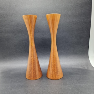 May include: Two wooden candle holders with a unique hourglass shape. The candle holders are made of light brown wood and have a smooth, polished finish. The design features a wide base that tapers to a narrow waist before widening again at the top.
