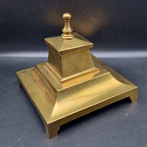 Puede incluir: Un accesorio de escritorio de latón con una base cuadrada y una estructura central escalonada rematada con un pequeño remate redondeado. La pieza tiene un acabado dorado pulido y es probablemente un portalápices o tintero.