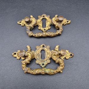 Könnte beinhalten: Zwei antike Schubladengriffe aus Messing mit Schlüssellochdetails. Jeder Griff weist verzierte Schnörkel und kleine Knöpfe auf beiden Seiten des Schlüssellochs auf. Das Messing hat ein Vintage-Aussehen, was darauf hindeutet, dass sie antik sind.