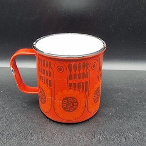 Puede incluir: Una taza vintage de esmalte con un exterior naranja vibrante y un interior blanco. La taza presenta un patrón repetido de pájaros estilizados y elementos florales en un tono más oscuro de naranja. El asa también es naranja.