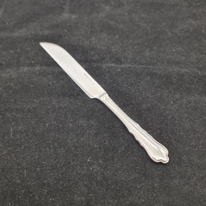 May include: A silver butter knife with a decorative handle. The knife has a slightly curved blade and a rounded tip.