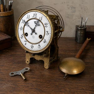 May include: An antique brass skeleton clock featuring a white circular dial with black Arabic numerals and ornate metal hands. The clock mechanism is exposed, showing internal gears and a coiled spring. A separate brass pendulum and winding key are included.