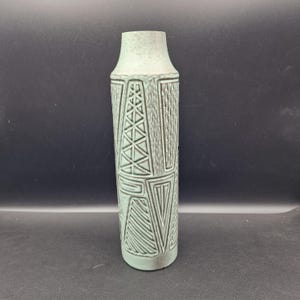 May include: A tall, light green vase with a textured surface and geometric patterns. The vase has a cylindrical body with a slightly flared top. The design includes triangles, lines, and rectangular shapes. The vase is made of ceramic or a similar material.