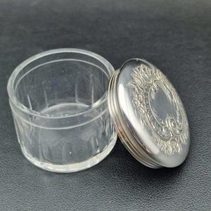 May include: A clear glass jar with a silver lid. The lid features an ornate floral design. The jar is open, showing its interior. The jar and lid are on a dark surface.