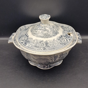 May include: A blue and white ceramic tureen with a lid, featuring a landscape and floral design. The tureen has handles and a spoon rest. The lid has a knob for easy removal. The tureen is a decorative serving dish.