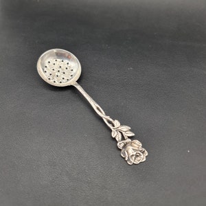 Puede incluir: Un colador de té de plata con un diseño de rosa. El colador tiene un cuenco redondo con agujeros y un mango en forma de tallo de rosa con hojas y un capullo de rosa.