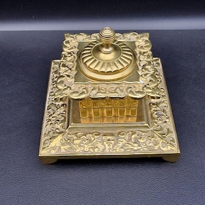 May include: Ornate antique brass inkwell with a square base and a hinged lid. The inkwell features detailed floral and scrollwork patterns, and a small, round finial on top. The overall design is symmetrical and decorative.