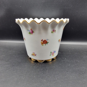 Peut inclure: Pot de fleurs en céramique blanche avec un bord festonné et une bordure dorée. Le pot est décoré de motifs floraux colorés. La base a également une bordure dorée. Une pièce décorative pour les plantes.