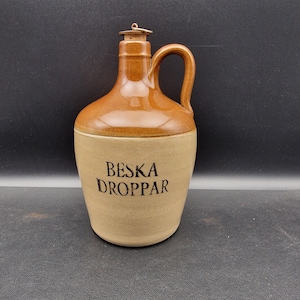 Può includere: Una brocca in ceramica marrone e beige con manico e tappo in sughero. La brocca ha le parole "BESKA DROPPAR" stampate sul davanti. La brocca è di colore beige chiaro nella parte inferiore e marrone più scuro nella parte superiore.