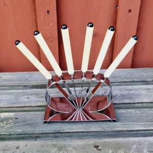 May include: A set of six silver cocktail picks with black and white handles, displayed in a silver stand. The stand is shaped like a fan and has a mirrored finish.