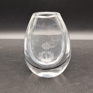 Könnte beinhalten: Klarer Glasvase mit ovaler Form und einem dekorativen Emblem aus Krone und Lorbeerblättern. Die Vase hat ein schwarzes Band in der Mitte und steht vor einem dunklen Hintergrund. Höhe ca. 10 cm.