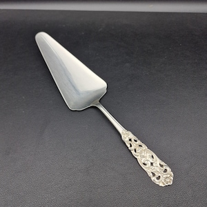May include: A silver cake server with a triangular blade and a decorative handle. The handle features an ornate floral and scroll design. The server is made of a shiny, reflective metal and is set against a dark background.