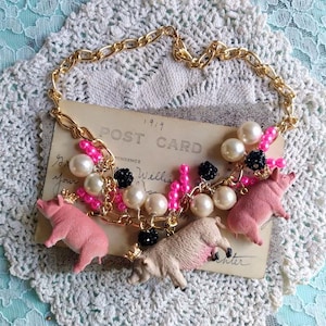 May include: A whimsical necklace featuring pink pig charms, faux pearls, and black bead accents. The gold-tone chain is adorned with pink beads. A vintage postcard is visible in the background.