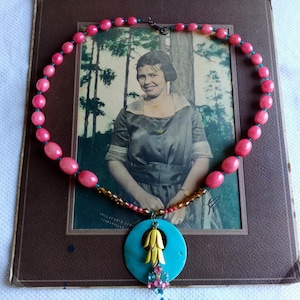 May include: A vintage necklace with pink beads and a turquoise pendant with yellow and blue accents. The necklace is displayed on a framed vintage photograph of a woman in a dress.