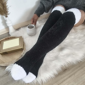May include: Black and white over-the-knee socks. The socks are black with white cuffs and toes. The socks are made of a soft, fuzzy material. The person is wearing a grey jumper and sitting on a white fluffy rug.