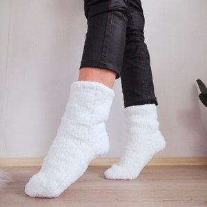 May include: A pair of white fuzzy socks with a ribbed texture. The socks are worn on a person's feet and legs. The person is wearing black pants.