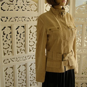 May include: A tan suede jacket with a zip fastening, a belt, and two large pockets on the front. The jacket has a stand-up collar and long sleeves.